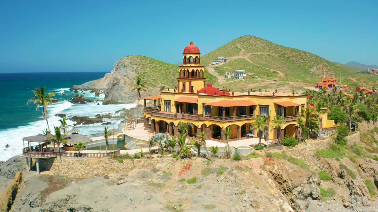 Wide aerial of church-like building, palms, cliffs, ocean, and hills.
