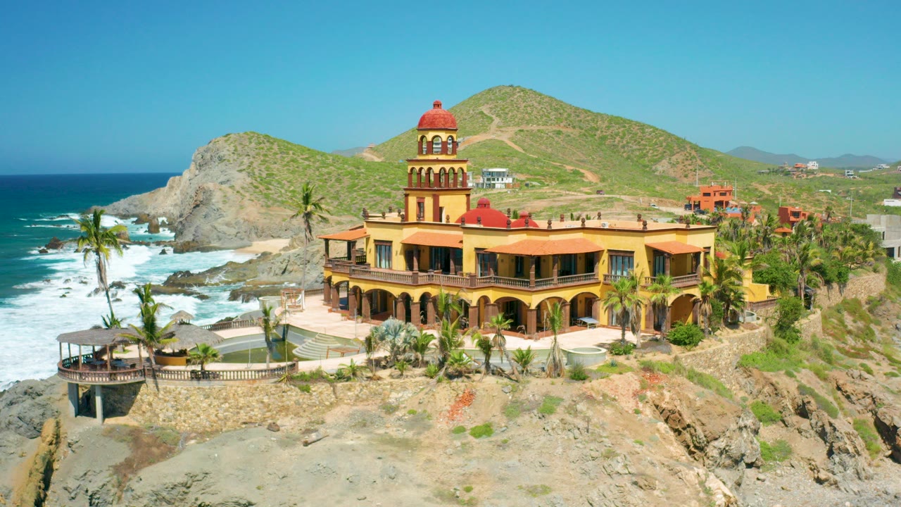 At 0:00, a yellow two-floor building with a red-domed tower overlooks rocky beach, palms, surf, and green hills in Cerritos, Baja California Sur, with no visible people.