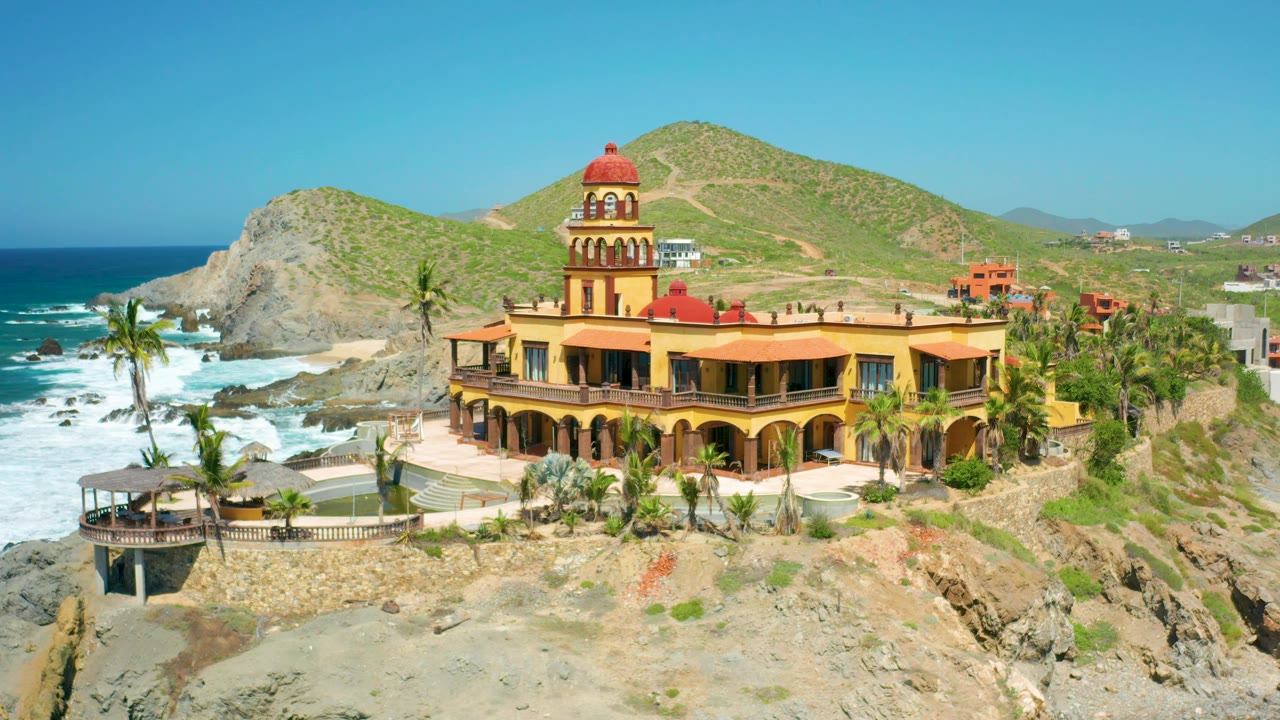 At 0:02, the elevated view shows the yellow property and tower slightly right of center, with surf and rock formations on the left and hillside buildings in the background, with no visible people.