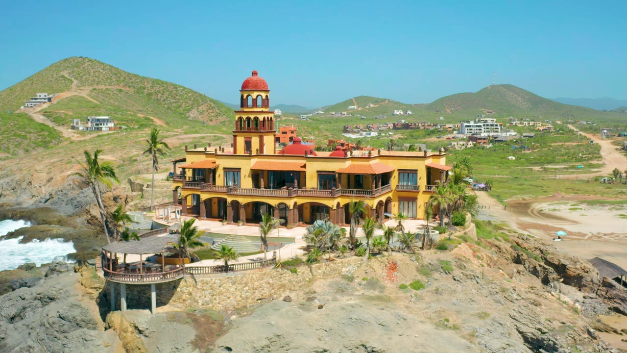 Aerial view of the castle-like building with surf, rocks, and inland hills.