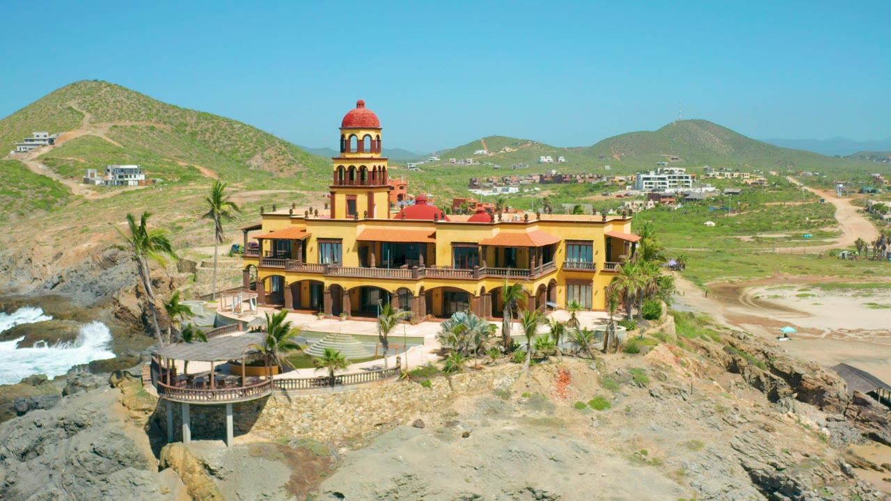 High daylight frame of the castle-like building in Cerritos, Baja California Sur, Mexico, with the terrace and palms visible in front, hills and scattered structures behind, and no visible people.