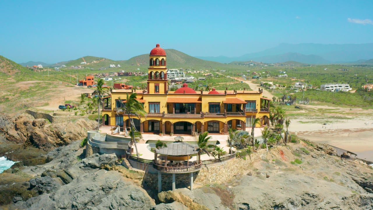 Aerial view in Cerritos, Baja California Sur of a yellow castle-like building with red domes above rocks and sand, with hills and no visible people.
