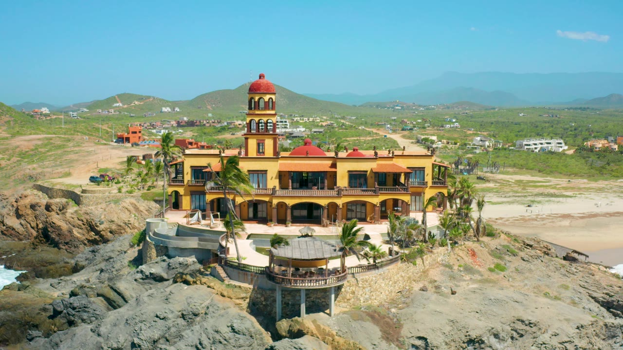 Aerial frame of the yellow castle-like structure in Cerritos with a red tower dome, rocky foreground, pale beach, inland hills, and no visible people.