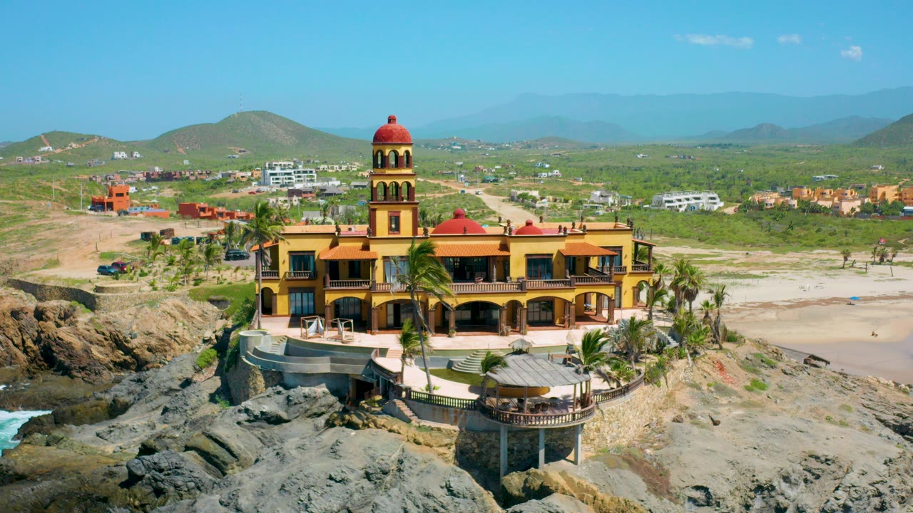 Aerial frame in Cerritos, Baja California Sur showing a yellow building with red domes beside dark rocks, pale sand, palms, inland scrub, and tiny visible people on the beach.
