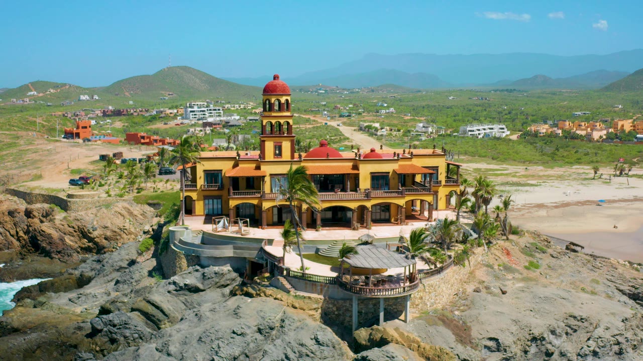 Aerial frame in Cerritos with the yellow and red domed building over rocks, a curved beach to the right, palms below, and hazy hills across the background.