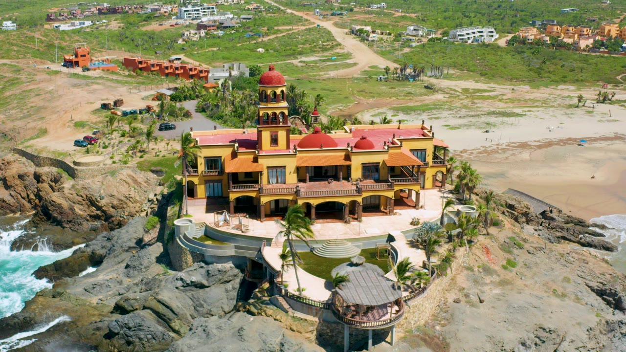 Aerial frame in Cerritos, Baja California Sur showing a yellow mansion with red domes on rocky shoreline, sandy beach and green scrub behind it, and no visible people.