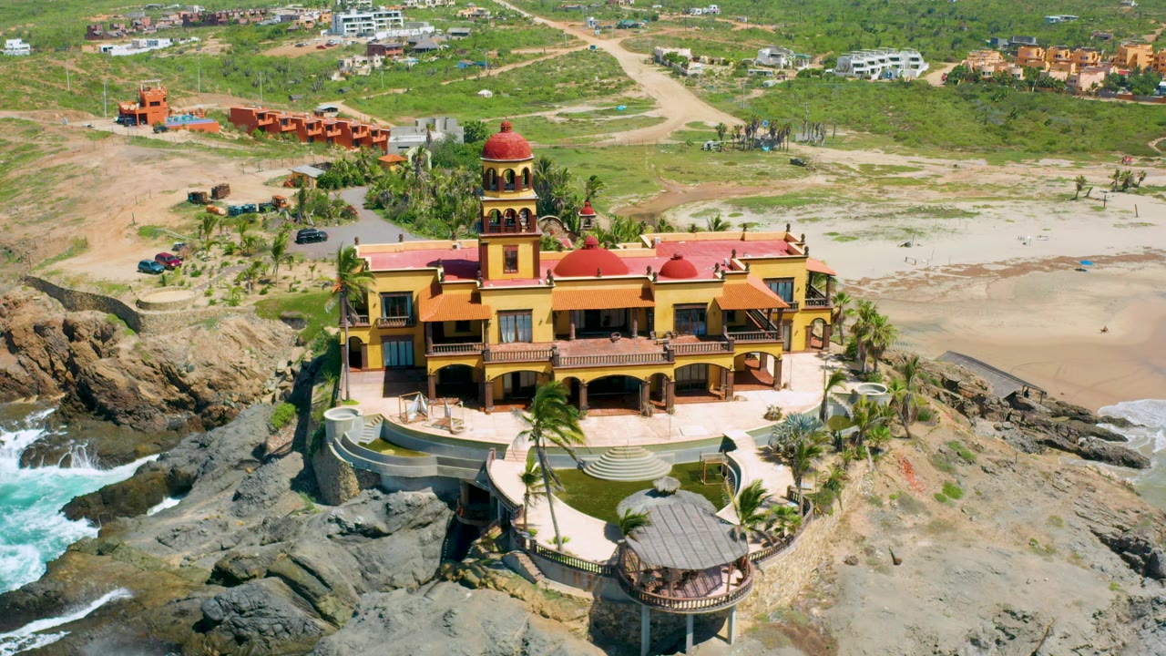 Drone view of yellow mansion beside rocks, beach, and surf.