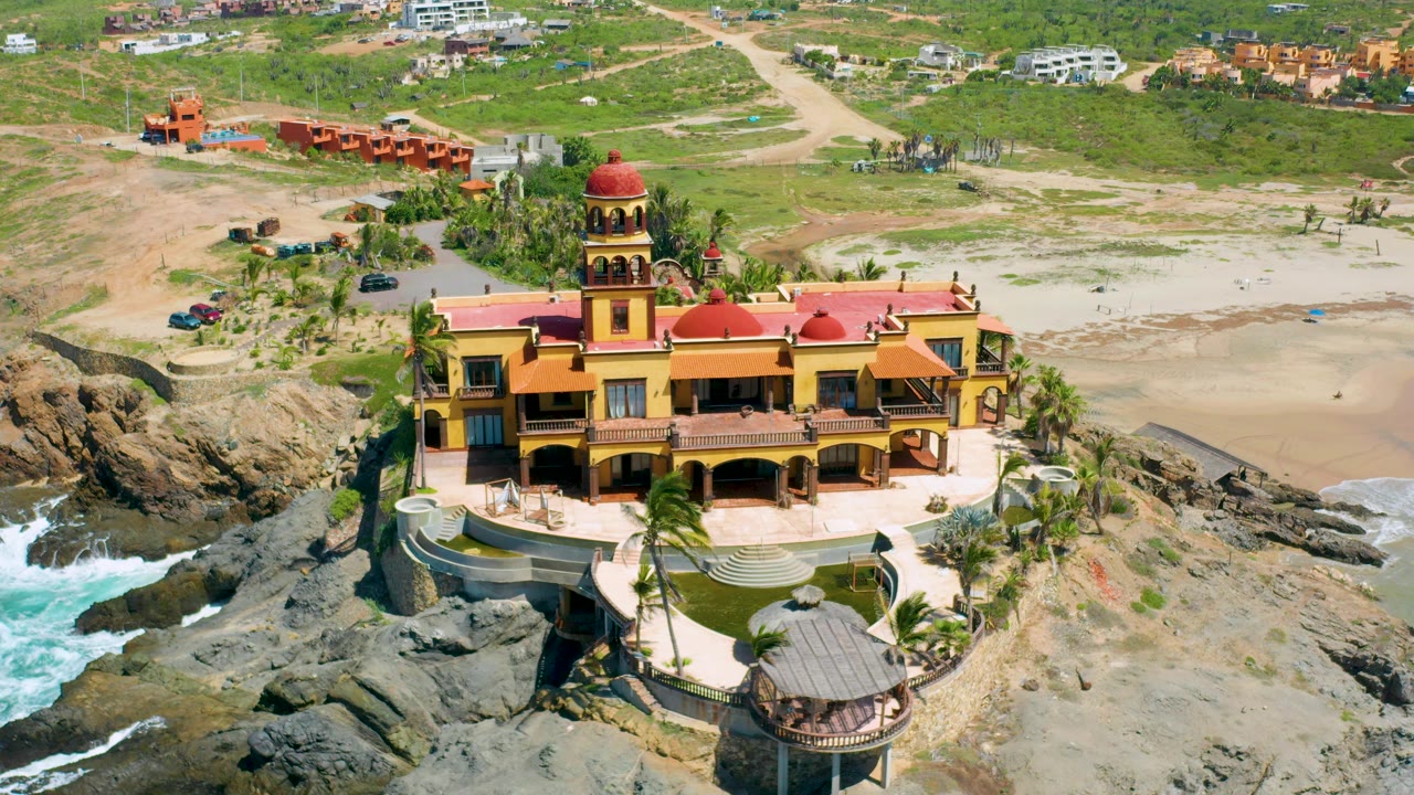 Cerritos, Baja California Sur aerial still showing the mansion on the rocky point, surf washing in at the left, sparse beachland behind it, and no visible people.