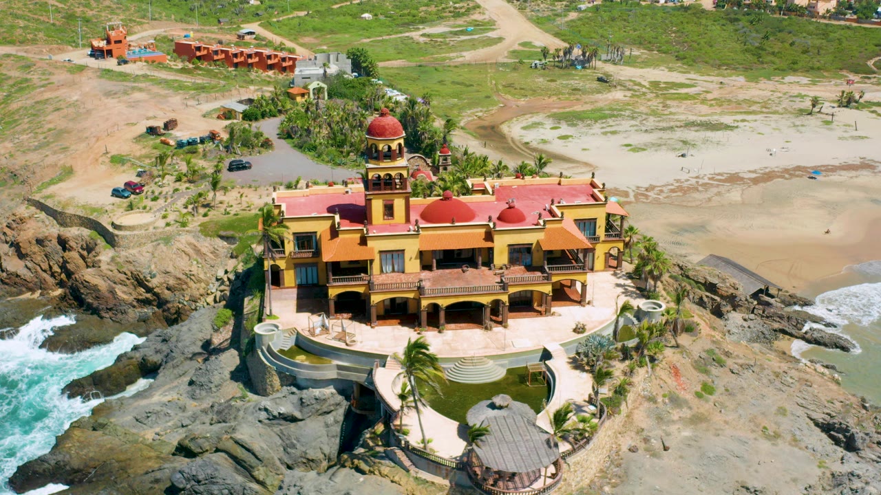 Aerial frame of a yellow and red mansion on the Cerritos, Baja California Sur shoreline, with dark rocks, white surf, beach sand, palms, and no visible people.