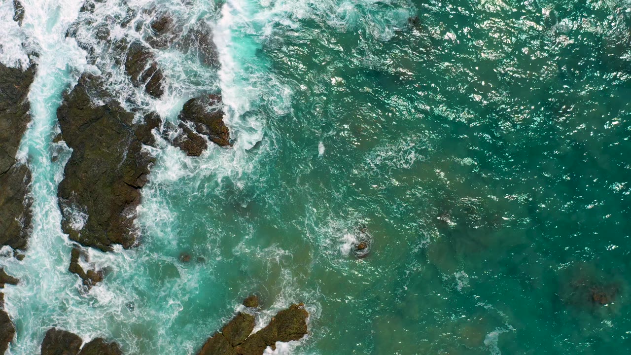 Foamy surf curls beside a long dark rock in clear green water.