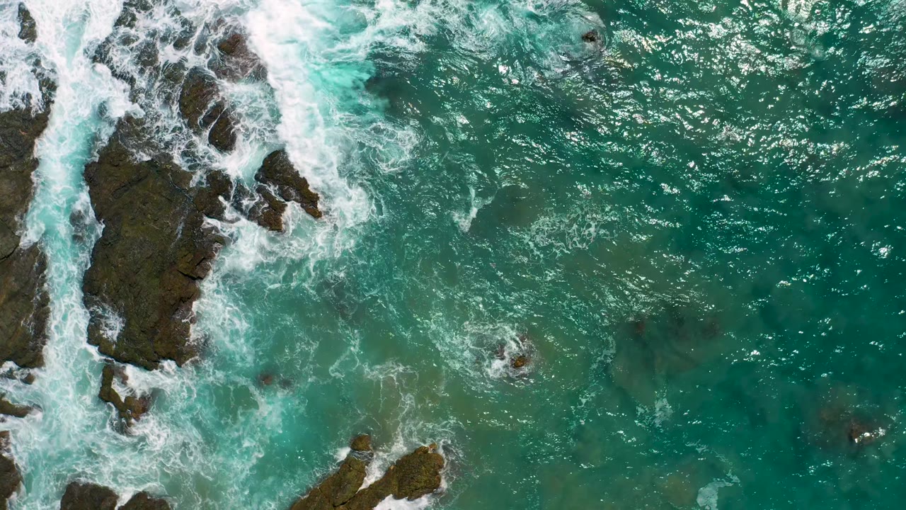 Overhead frame in Cerritos, Baja California Sur where white water spreads over the left-side rocks while open turquoise ocean fills the right half, with no visible people.