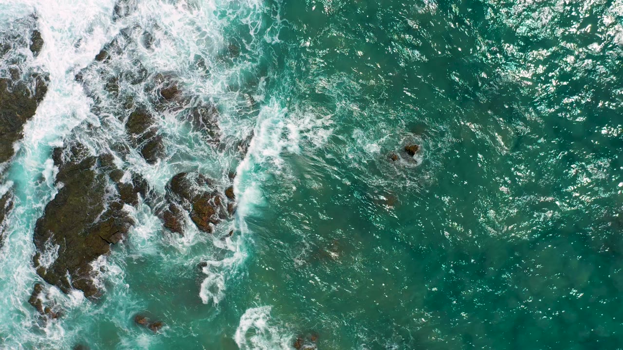 In Cerritos, Baja California Sur, white surf spreads across a central dark rock formation while clear blue-green water fills the surrounding frame, with no visible people.