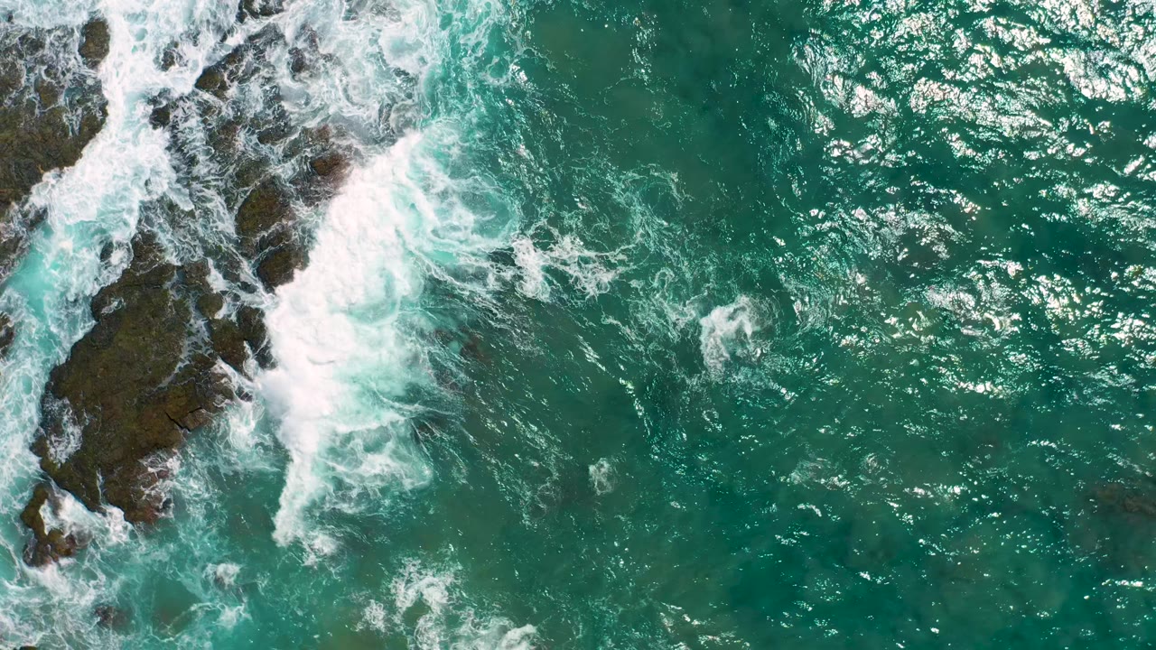 A Cerritos overhead frame shows heavy white wash pushing around rocks along the left edge, with calmer green-blue ocean and scattered sun highlights to the right, and no visible people.