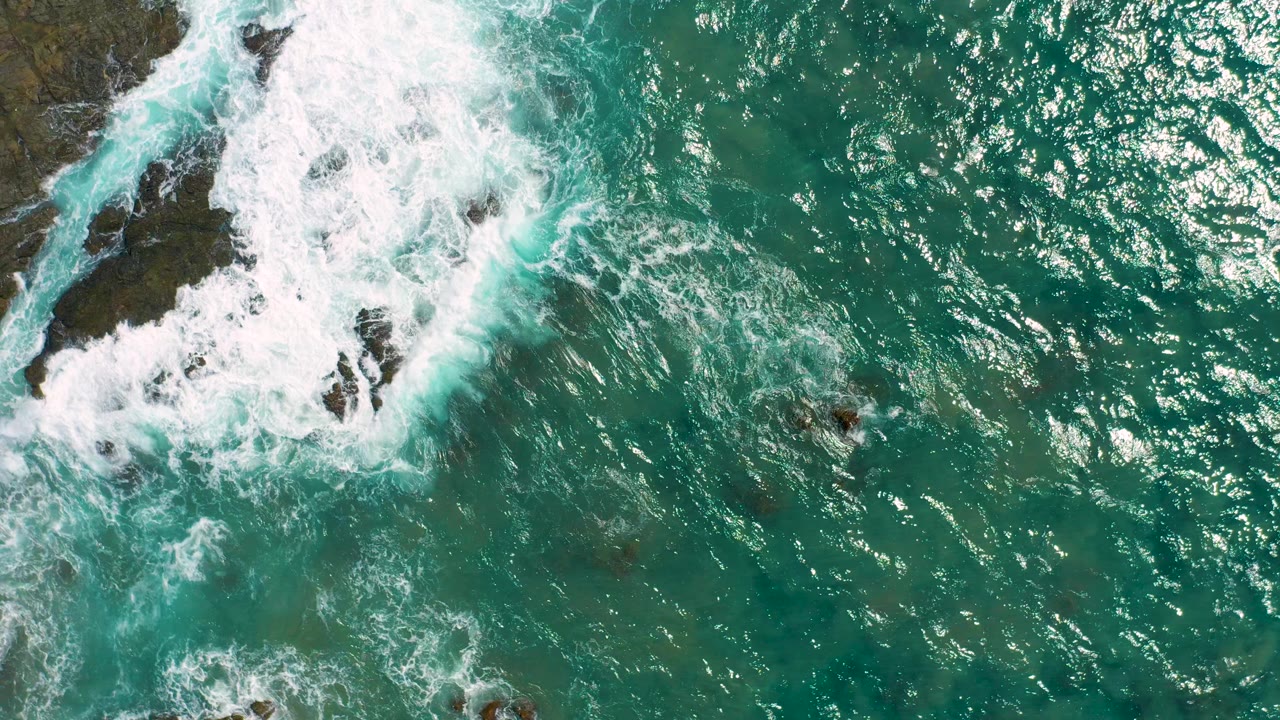 Overhead view of Cerritos, Baja California Sur surf washing around dark rock formations in turquoise water, with no visible people.