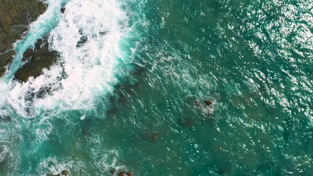 Top-down Cerritos coastal view with white surf covering upper-frame rocks and textured turquoise water extending across the right side, with no visible people.