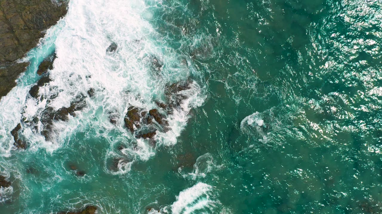 White foam spreads over rocks beside sunlit green-blue water.