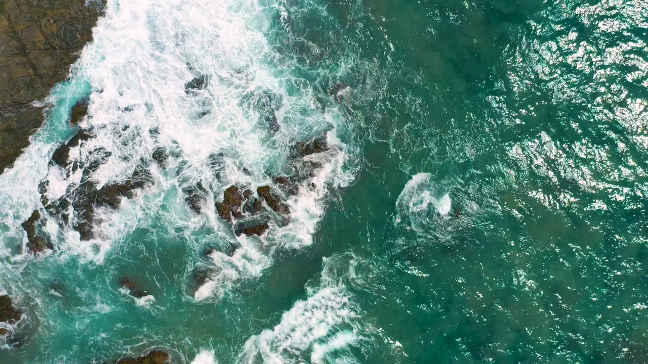 Surf curls around exposed dark rocks while rippled turquoise water occupies the right side of the Cerritos coastal frame, with no visible people.