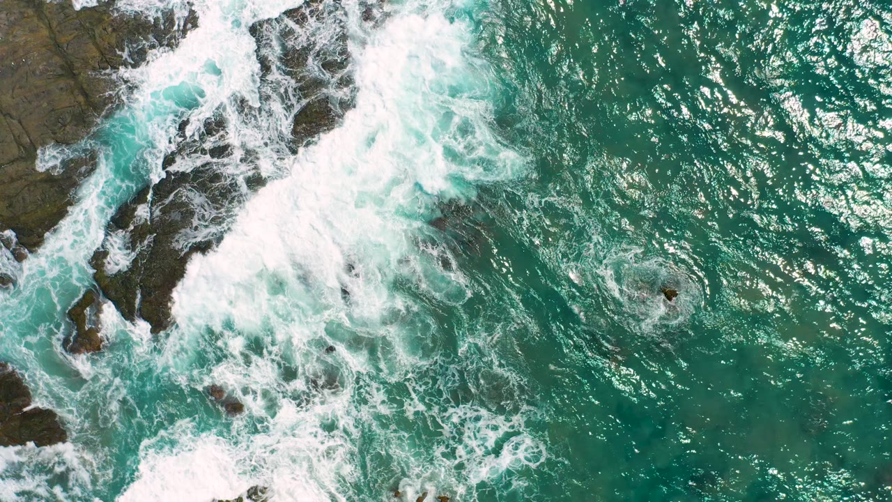 A top-down Cerritos shoreline frame shows white water curling around exposed dark rocks while green-blue ocean fills the right side, with no visible people.