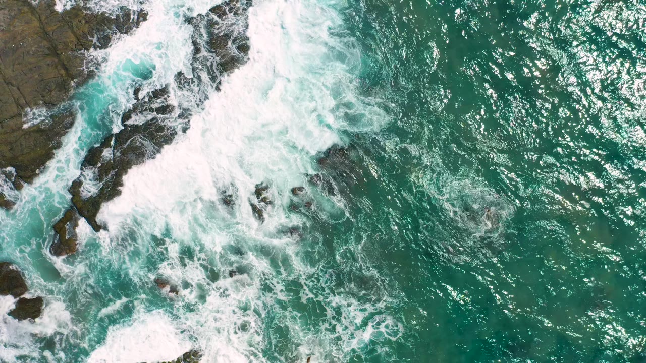 White foam fans out from the rocky left edge into turquoise ocean near Cerritos, with bright surface reflections and no visible people.