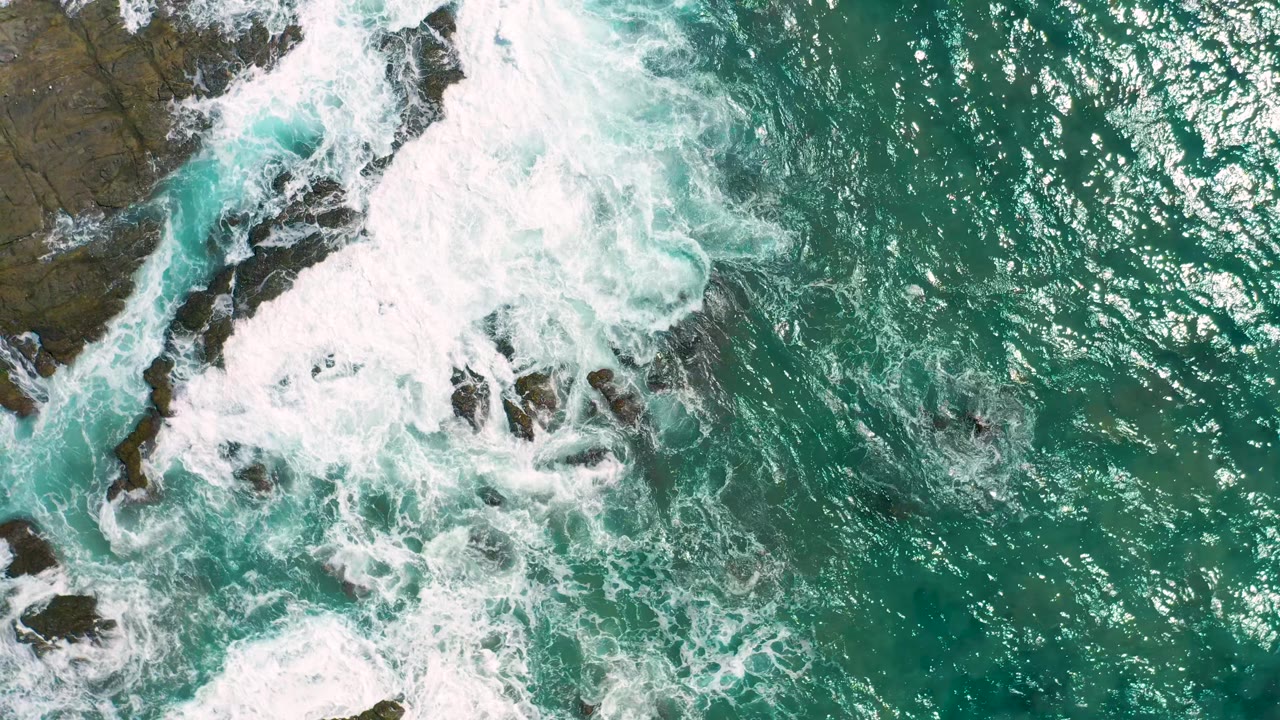 A large white patch of breaking surf covers dark rocks at the left while open teal water fills the right, with no visible people in the Cerritos frame.