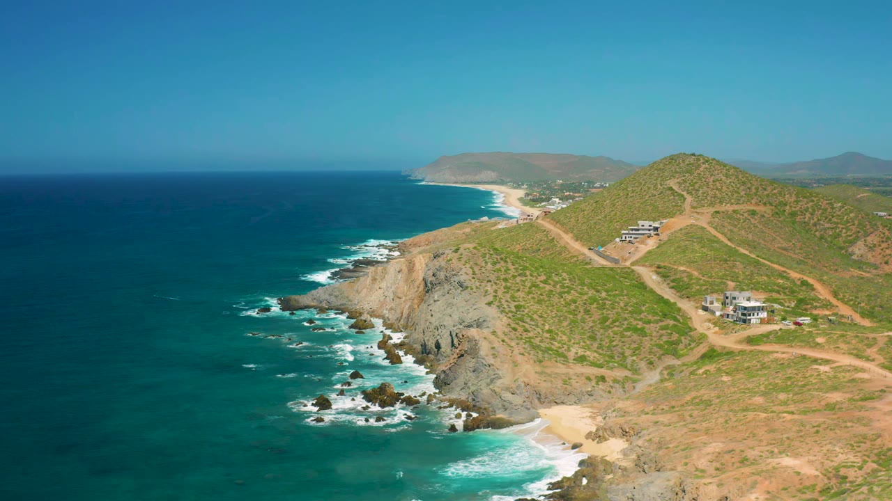Drone view of ocean surf beside cliffs and hillside buildings under a clear sky.