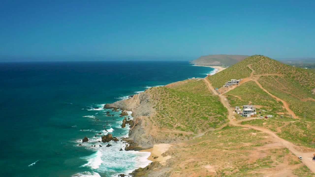A second aerial view in Cerritos, Baja California Sur keeps the ocean broad on the left while surf curls into the rocks below hillside houses, with no visible people.
