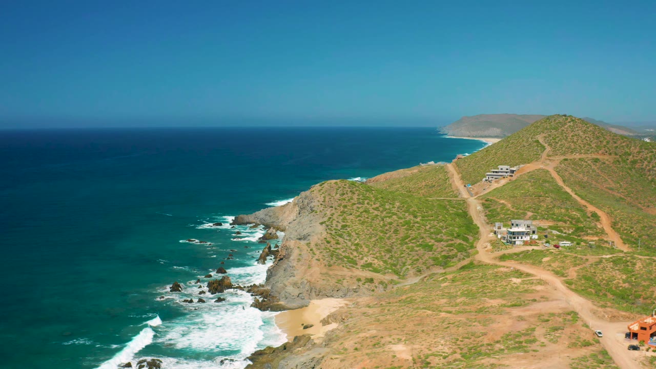 Aerial frame over Cerritos, Baja California Sur showing the cliff edge closer to center, deep blue water to the left, hillside tracks to the right, small buildings, and no visible people.