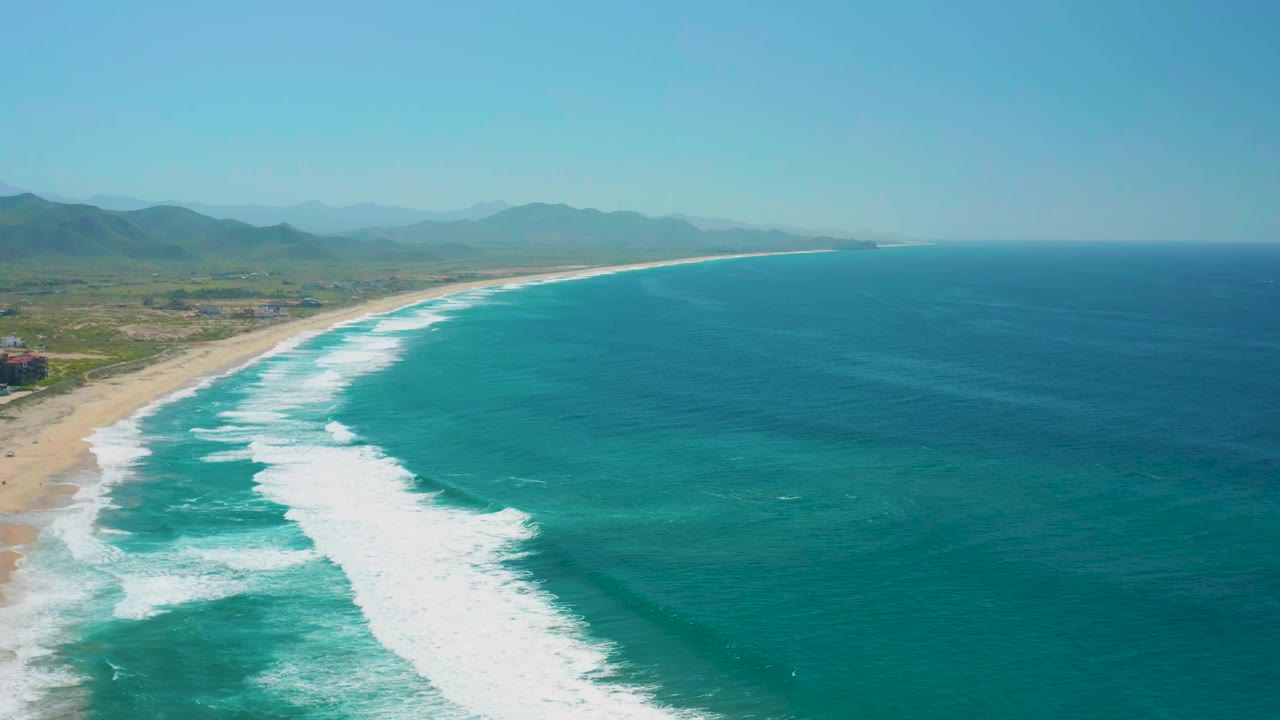 Aerial view of Cerritos coastline with the surf band running diagonally through the frame, open blue water to the right, hazy hills inland, and no visible people.