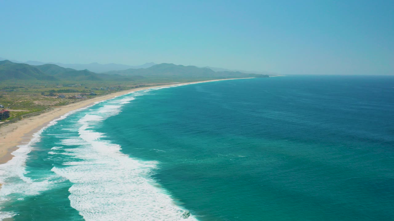 Aerial frame over Cerritos, Baja California Sur showing white surf bands meeting a sandy shore, turquoise ocean to the right, green land and hazy mountains inland, with no visible people.