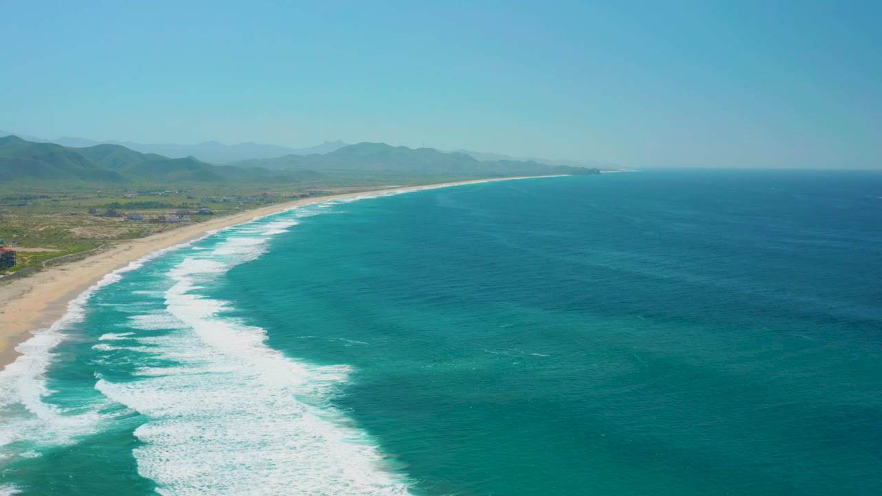 Cerritos beach appears from above with the wave break shifted along the shore, turquoise shallows near the surf, darker ocean farther right, green land to the left, and no visible people.