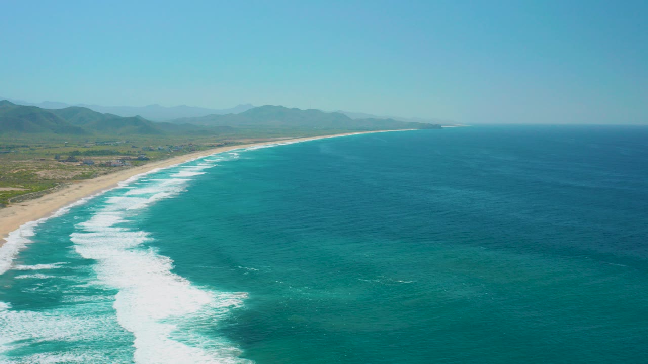 Aerial frame over Cerritos, Baja California Sur showing turquoise water, a broad line of white surf, sandy beach at left, distant low hills, and no visible people.