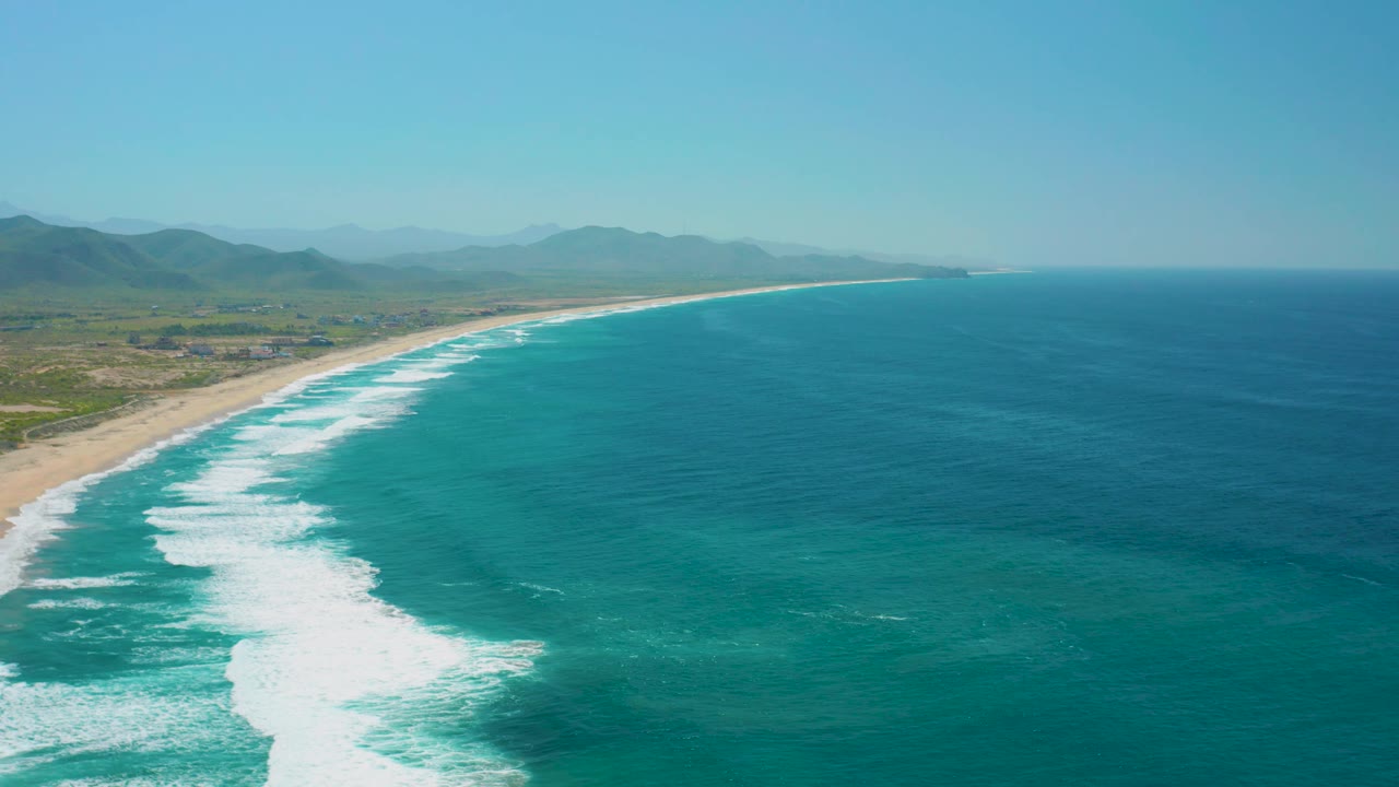 At 0:02, the aerial view shows a slightly longer reach of Cerritos shoreline, with the surf line curving toward the horizon, inland flats at left, and no visible people.