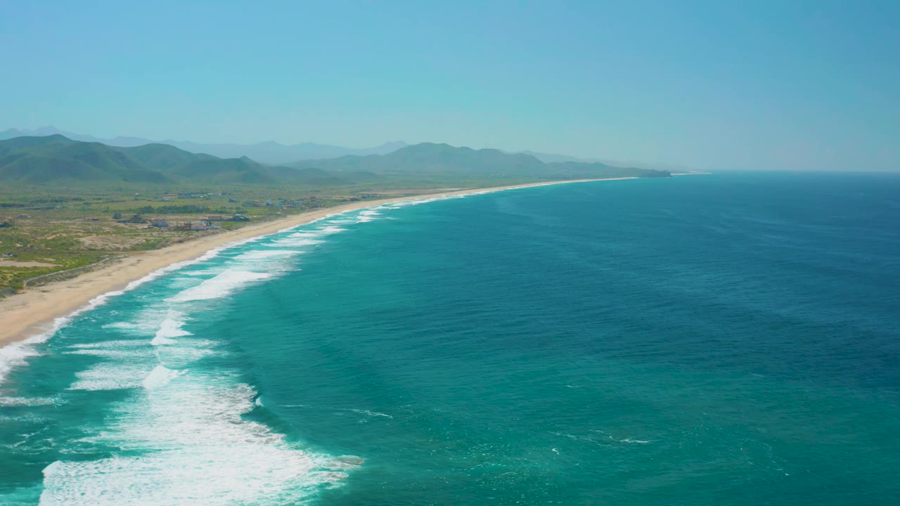 High aerial frame at Cerritos showing white surf bands pushing toward the sandy beach, broad blue-green ocean on the right, hazy mountains inland, and no visible people.
