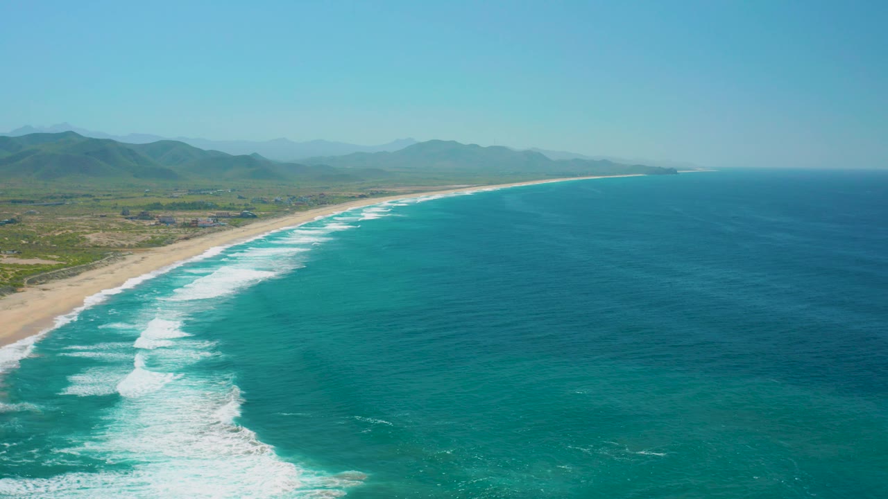 Wide aerial frame over Cerritos with surf entering from the lower left, a pale sandy beach running toward the horizon, open ocean to the right, inland mountains, and no visible people.