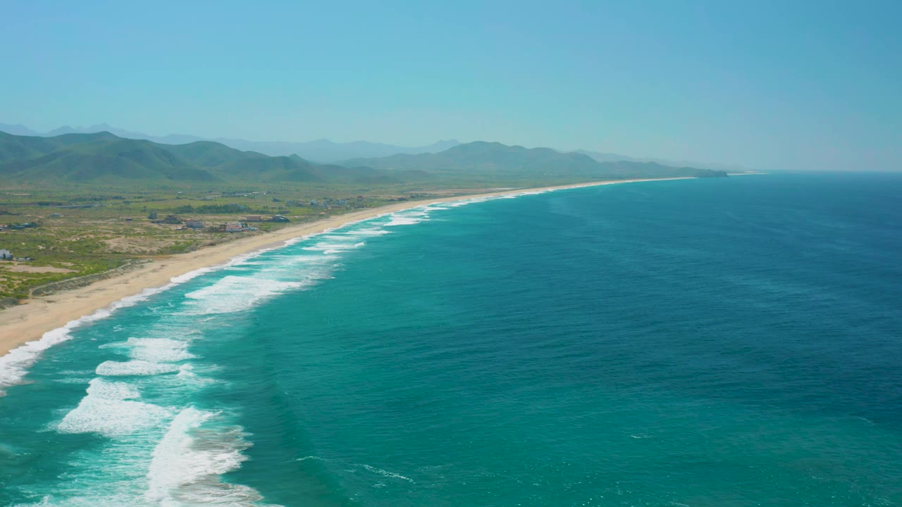 Aerial coastline with breaking waves, sandy beach, inland land, and mountains.