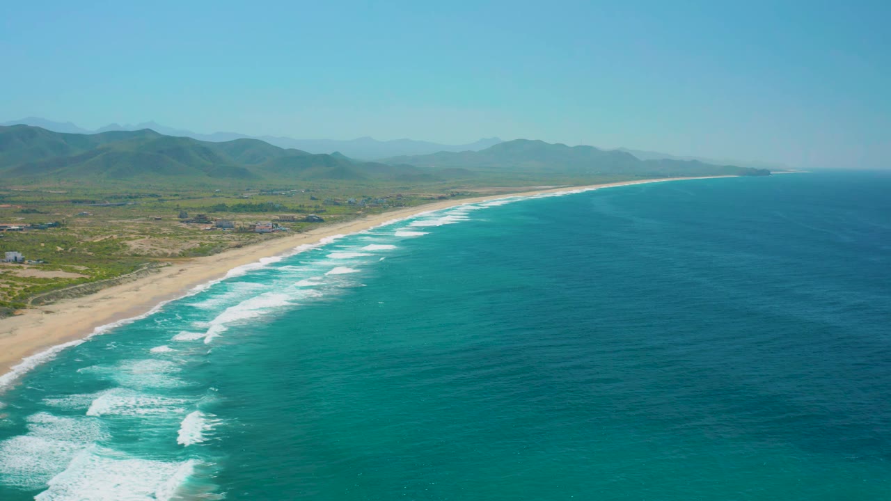 Wide aerial frame from Cerritos, Baja California Sur, showing foamy waves rolling toward the beach, blue-green ocean, open coastal land, hazy mountain ridges, and no visible people.