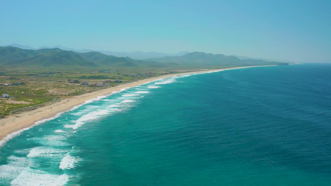 Drone-height view over Cerritos, Baja California Sur, with the long beach arc cutting between pale sand and deep blue water, surf clustered near shore, low inland scrub, mountains, and no visible people.