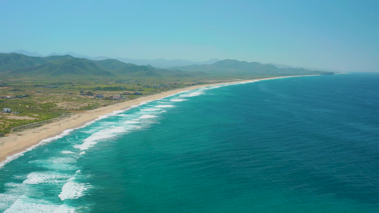 Aerial view of Cerritos, Baja California Sur, where white surf traces a sandy coastline, the inland side shows open terrain and scattered small structures, distant mountains sit under clear sky, and no visible people appear.