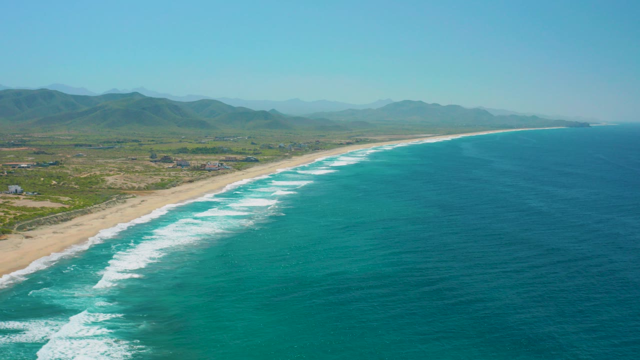 A high aerial frame over Cerritos, Baja California Sur shows turquoise water, white surf bands, a sandy shoreline, green coastal land, distant mountains, and no visible people.