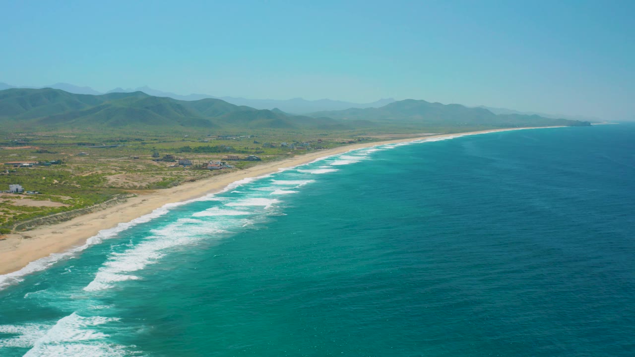At 0:02, the aerial view favors the open ocean on the right while the Cerritos beach curves toward distant headland shapes, with inland mountains and no visible people.