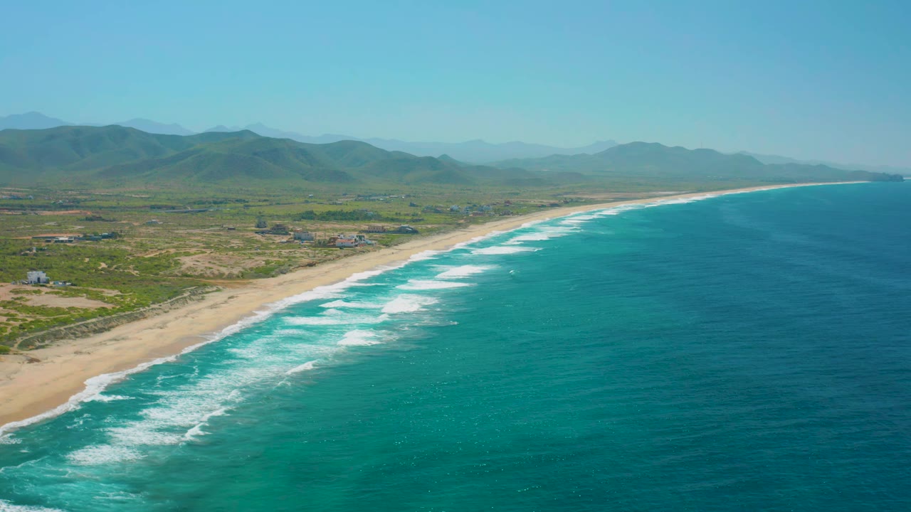 Wide aerial coastline with white wave bands rolling toward sand and mountains beyond.