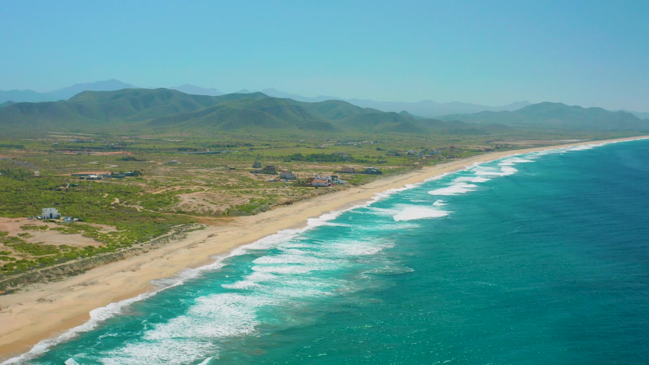 Aerial view of Cerritos, Baja California Sur, with turquoise ocean on the right, white surf bands along the sand, green coastal terrain, distant mountains, and no visible people.
