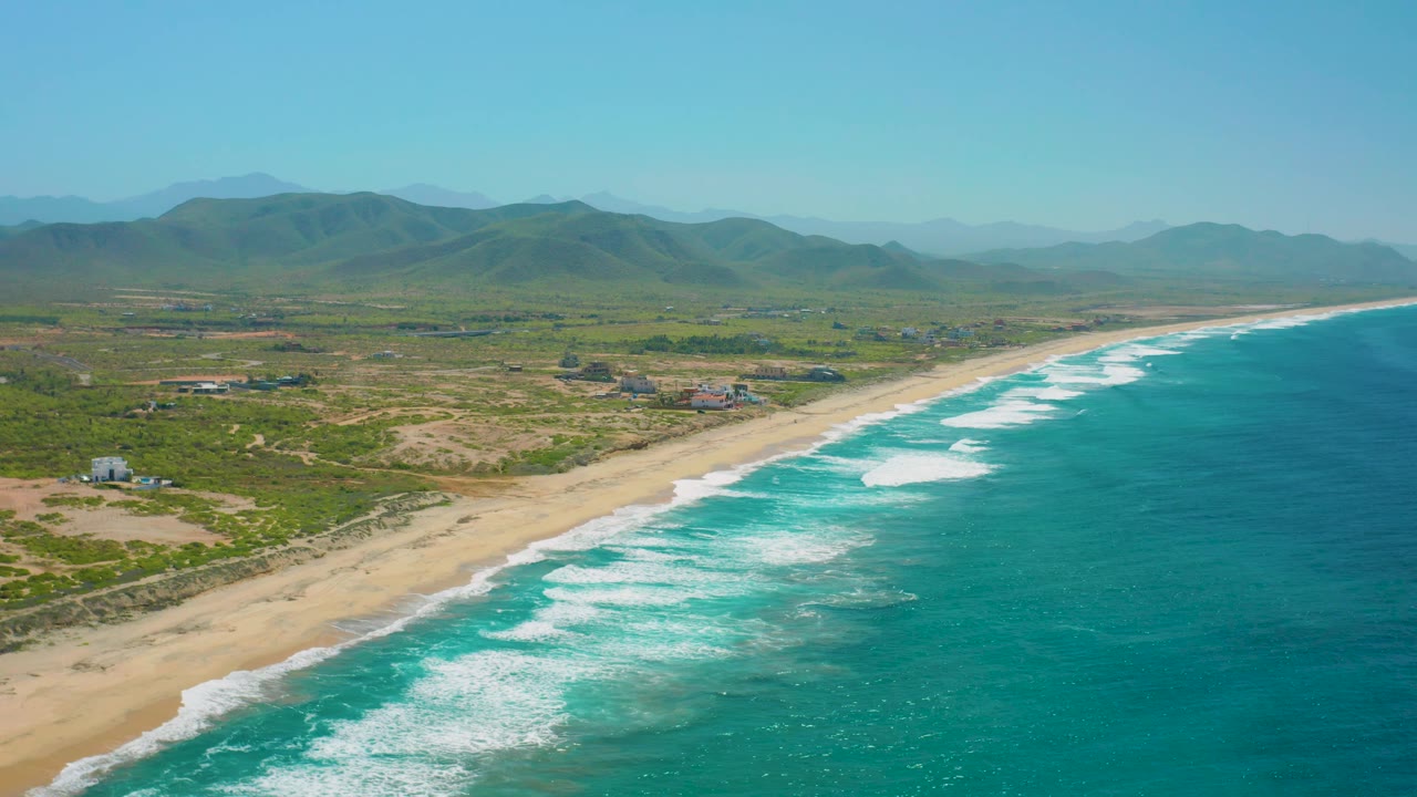 Aerial Cerritos coastline frame with ocean occupying the right half, foam lines reaching the beach, low green terrain with scattered structures, mountains, and no visible people.