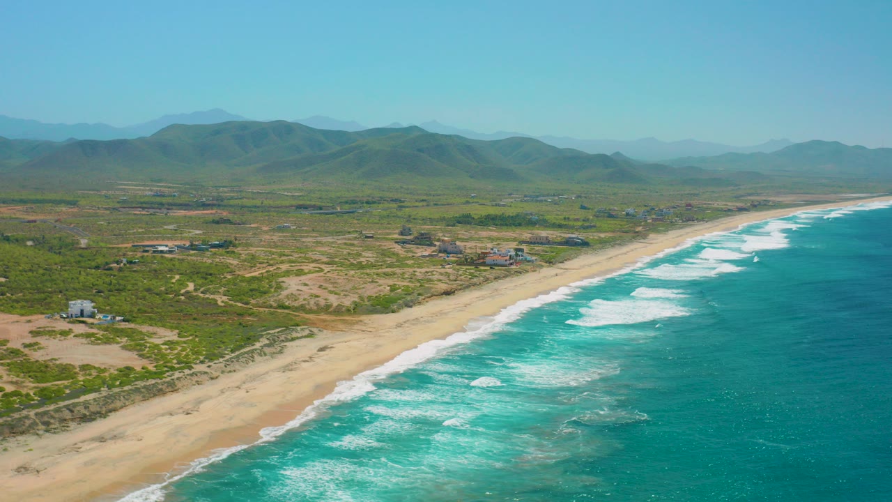 Opening aerial frame over Cerritos, Baja California Sur, with turquoise waves on the right, a diagonal sandy beach, inland vegetation, scattered buildings, distant hills, and no visible people.