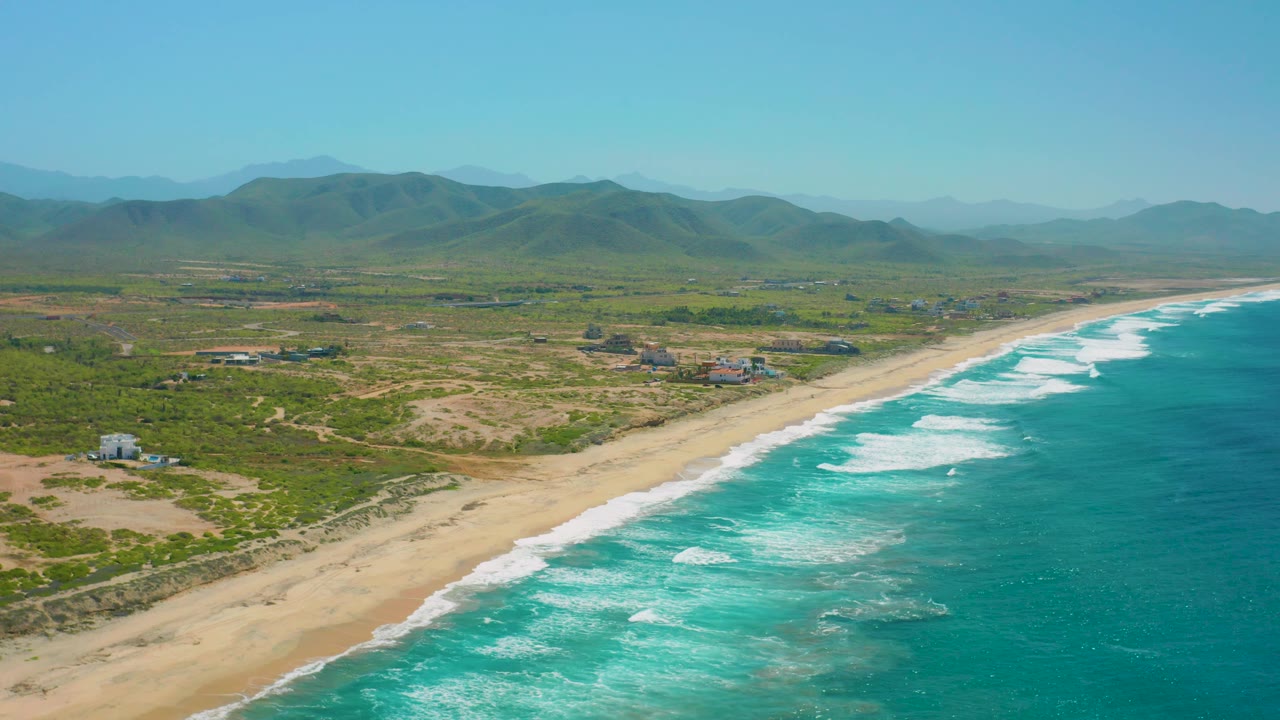 Cerritos Beach appears from above with bright blue water, white wave bands, tan sand, patchy inland lots, a few structures, distant green hills, and no visible people.