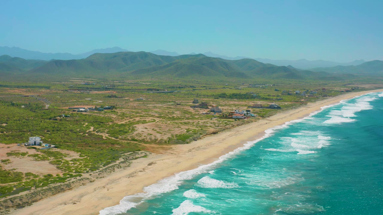 Wide coastal aerial with waves, beach, scrubland, and mountain ridge