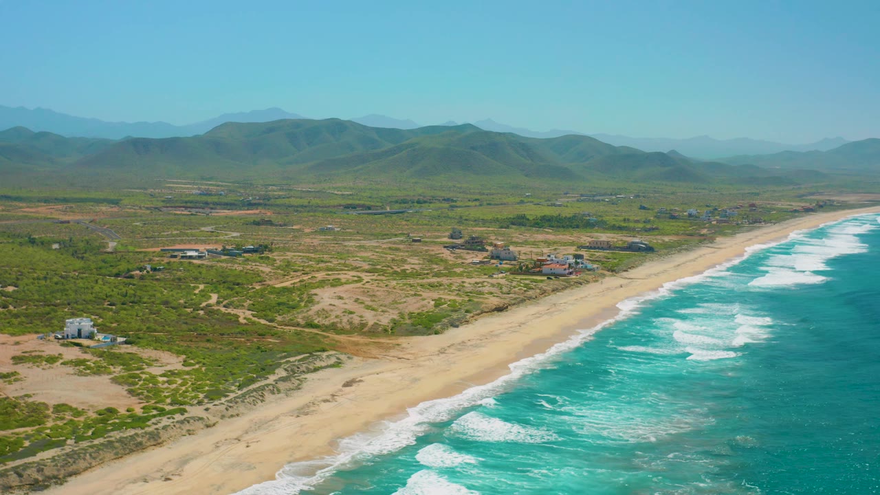 High aerial frame from Cerritos, Baja California Sur, showing ocean and surf on the right, a long pale beach, scrubby dunes, scattered inland buildings, hazy mountains, and no visible people.