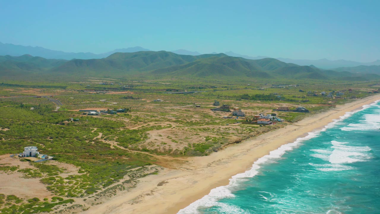 Cerritos coastline from above with white wave lines closer to the lower right, open tan beach through the center, low vegetation inland, small buildings, mountains, and no visible people.