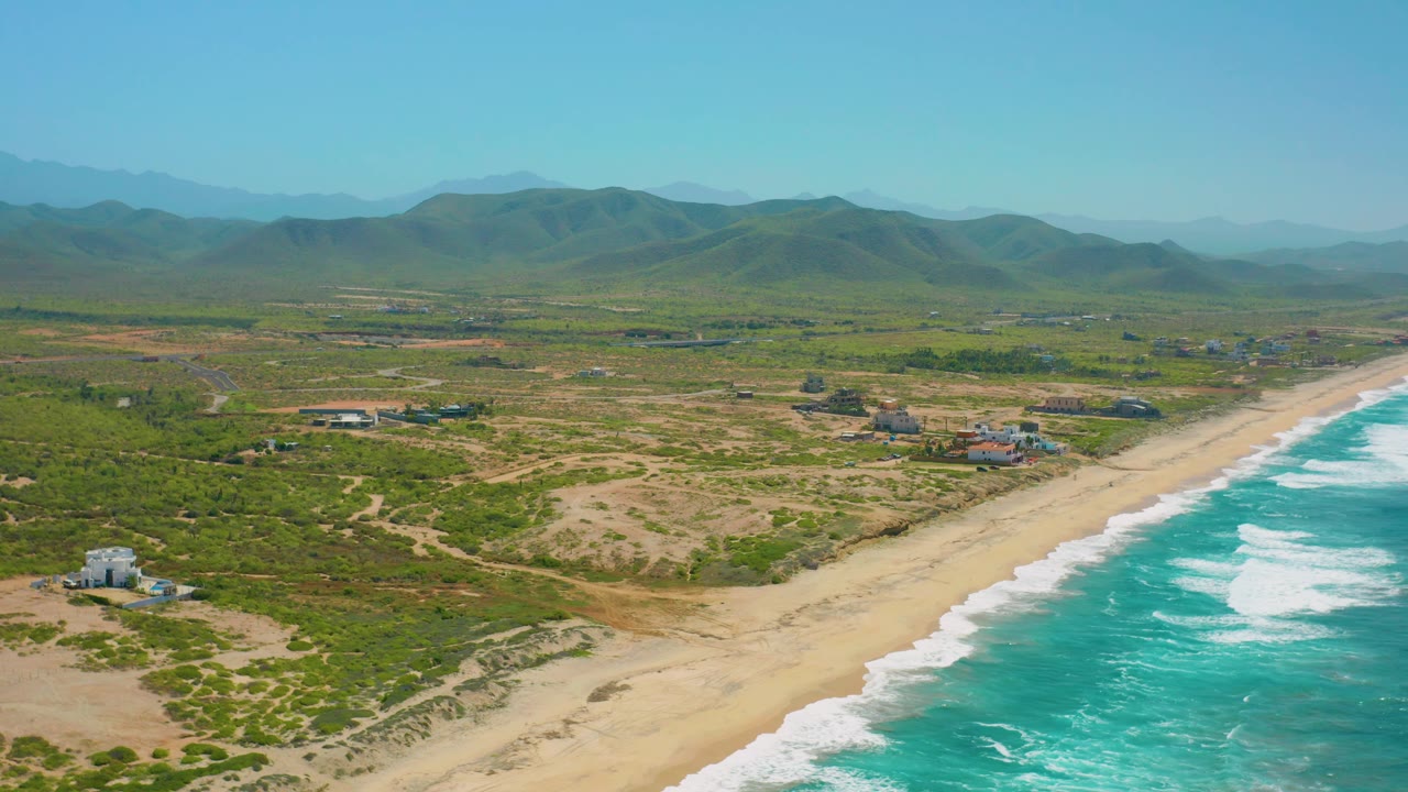 High view over Cerritos beach with foamy turquoise water on the right, uninterrupted sand below the dunes, scattered homes inland, hazy mountain layers, and no visible people.