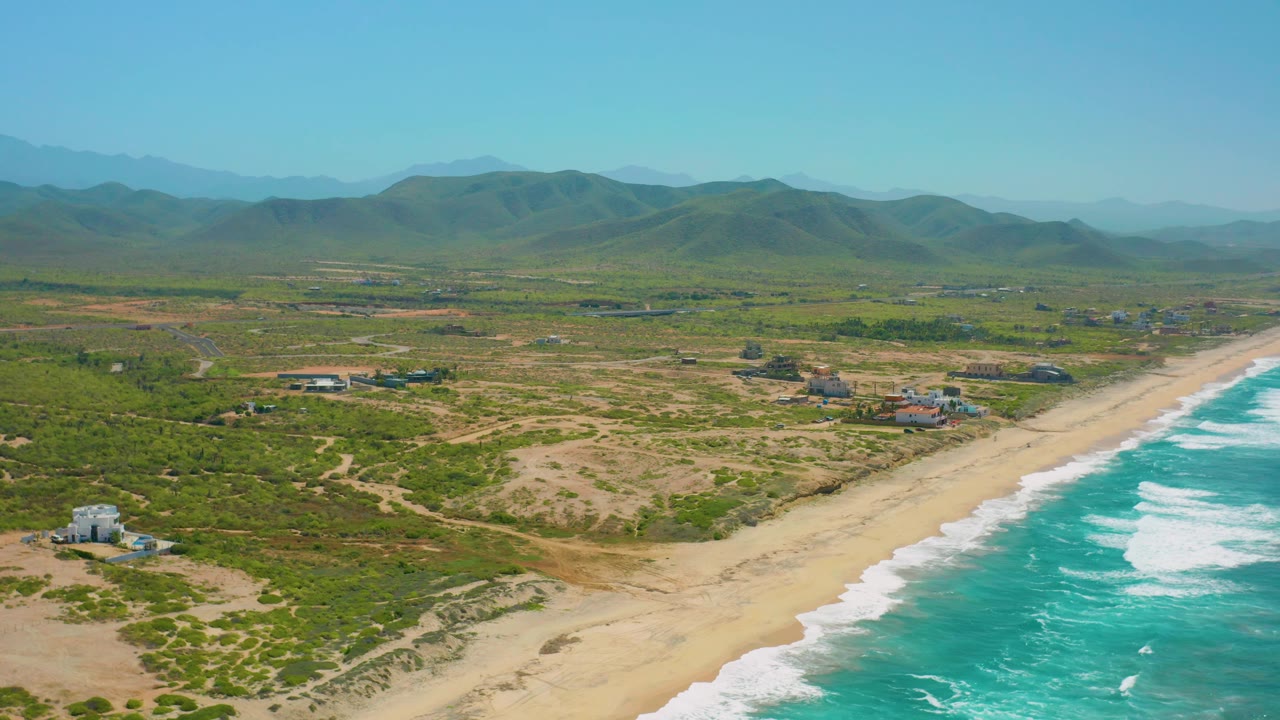 Aerial view of a quiet beach, surf, scrubland, buildings, and mountains.